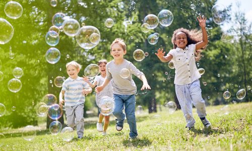 Multi-ethnic group of little friends with toothy smiles on their faces enjoying warm sunny day while participating in soap bubbles show